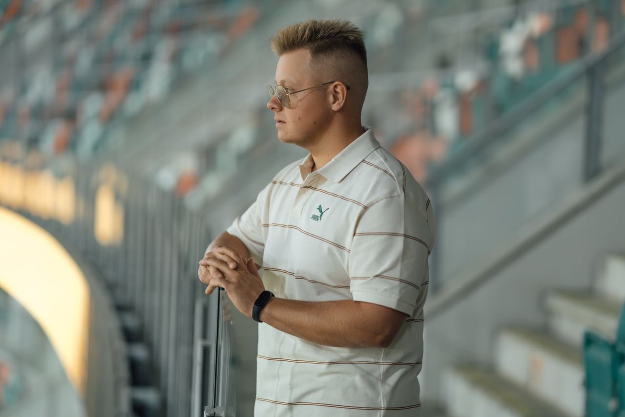 about-img Man standing thoughtfully in a stadium, wearing a striped polo shirt and glasses.